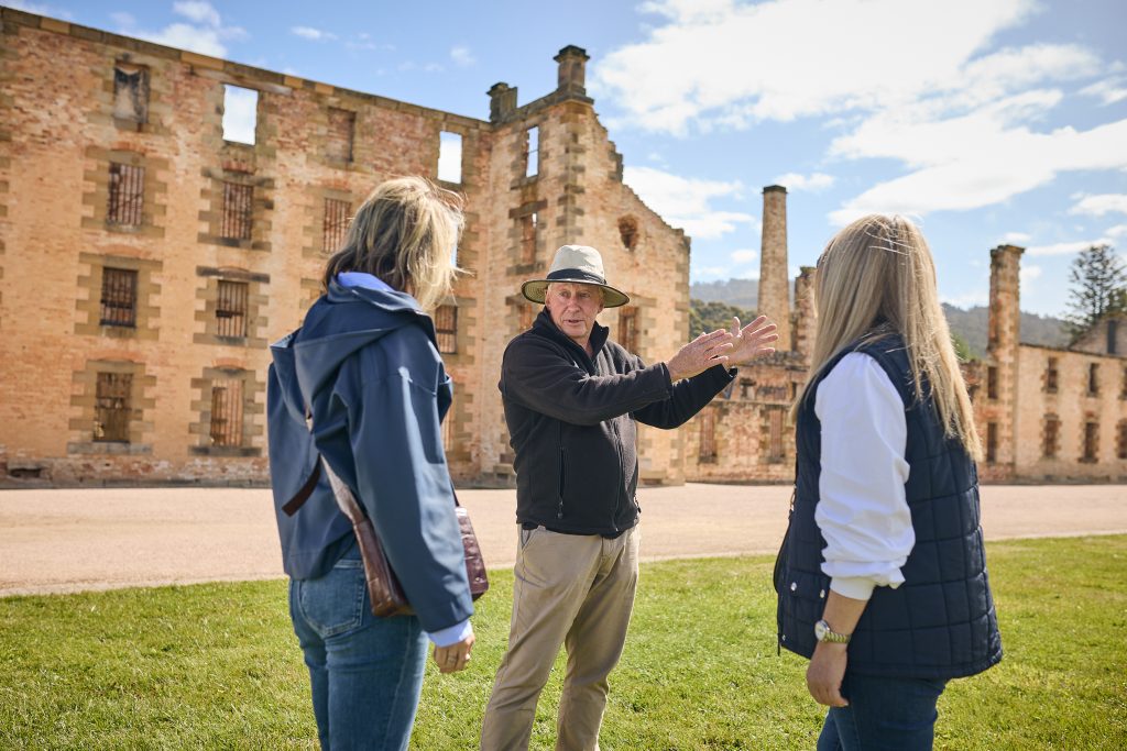 A Port Arthur Guide in uniform gestures towards the ruins of the Penitentiary watched in interest by two visitors
