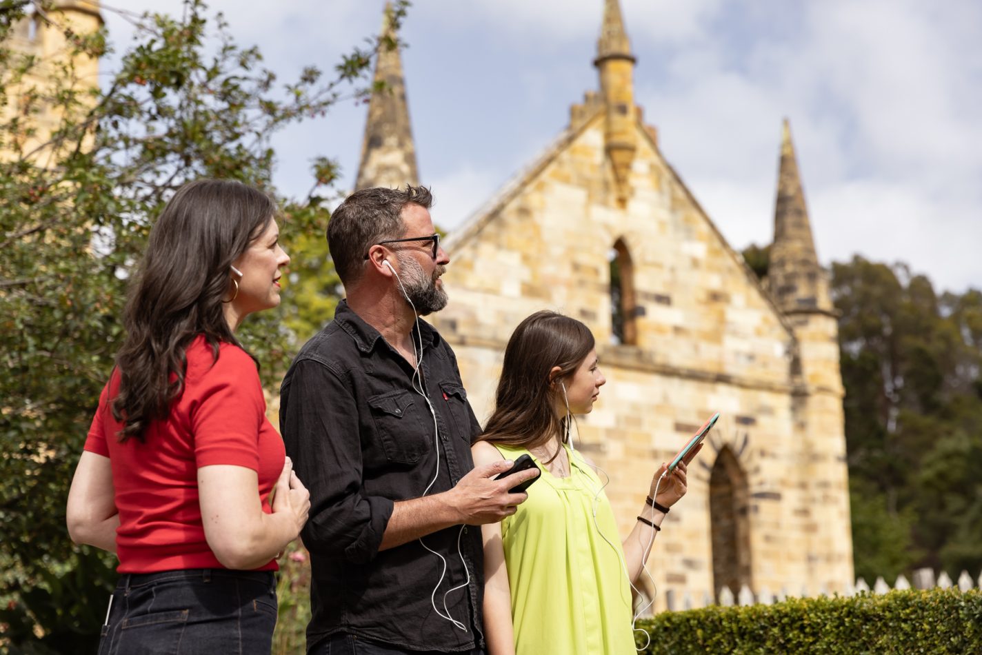 A mother, father and daughter listen to the audio experience on their phones with earbuds in front of the Port Arthur Church
