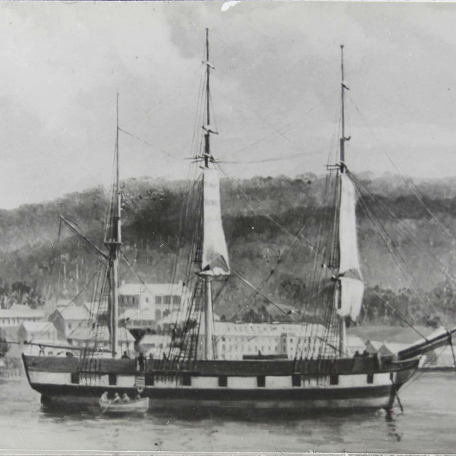 Historic black and white photograph of a sailing ship anchored near a coastal town with hills in the background.