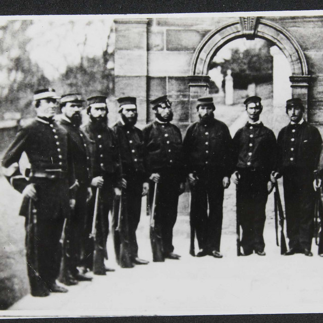 Vintage photograph of eight uniformed men standing in a line, likely 19th century soldiers or police officers.