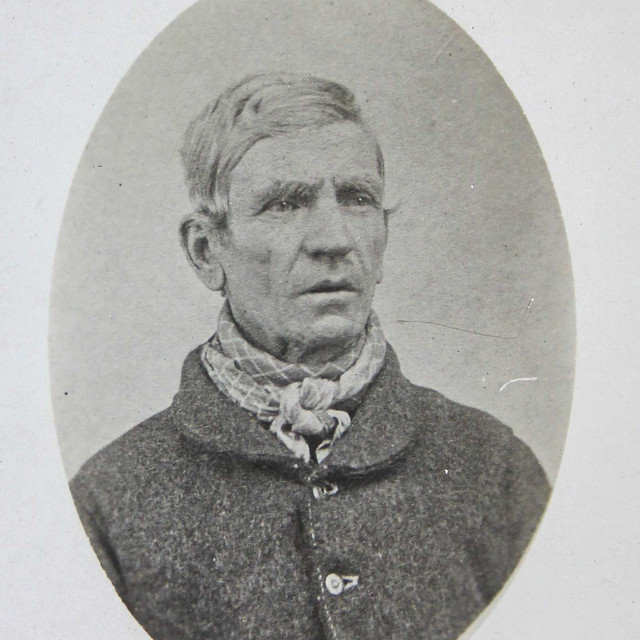 Vintage black and white portrait of an older man with short hair, wearing a dark coat and patterned neckerchief.