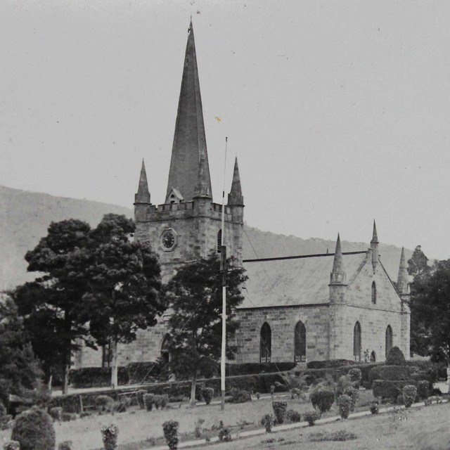 A historic church with a tall spire, surrounded by neatly trimmed shrubs and trees. A mountain range is visible in the background.