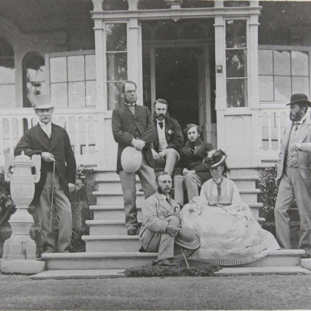 A group of people in 19th-century attire pose on the steps of an ornate building, with one woman sitting and five men standing around her.